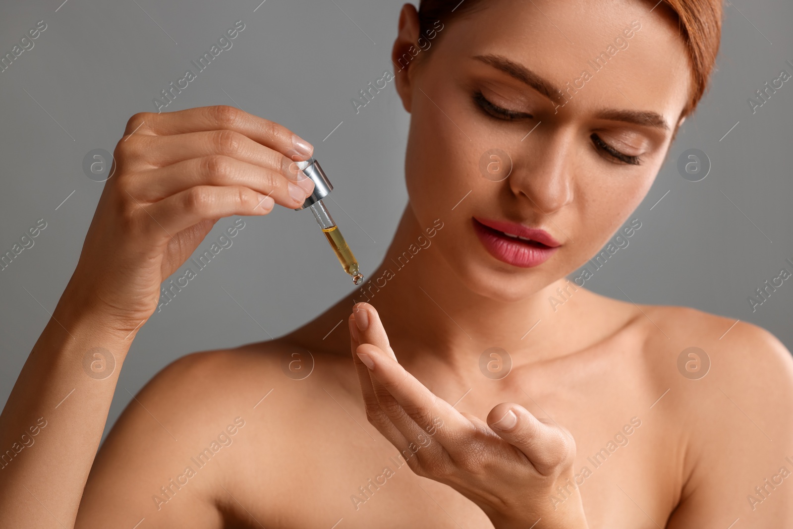 Photo of Beautiful young woman applying cosmetic serum onto her finger on grey background, closeup