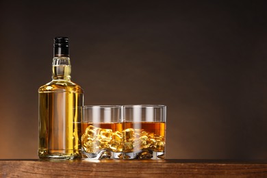 Photo of Whiskey with ice cubes in glasses and bottle on wooden table against brown background, space for text