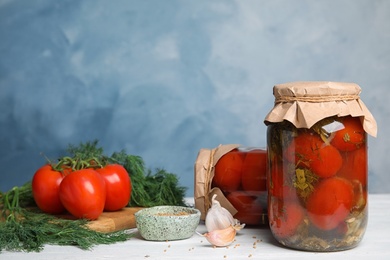 Pickled tomatoes in glass jars and products on white wooden table against blue background, space for text