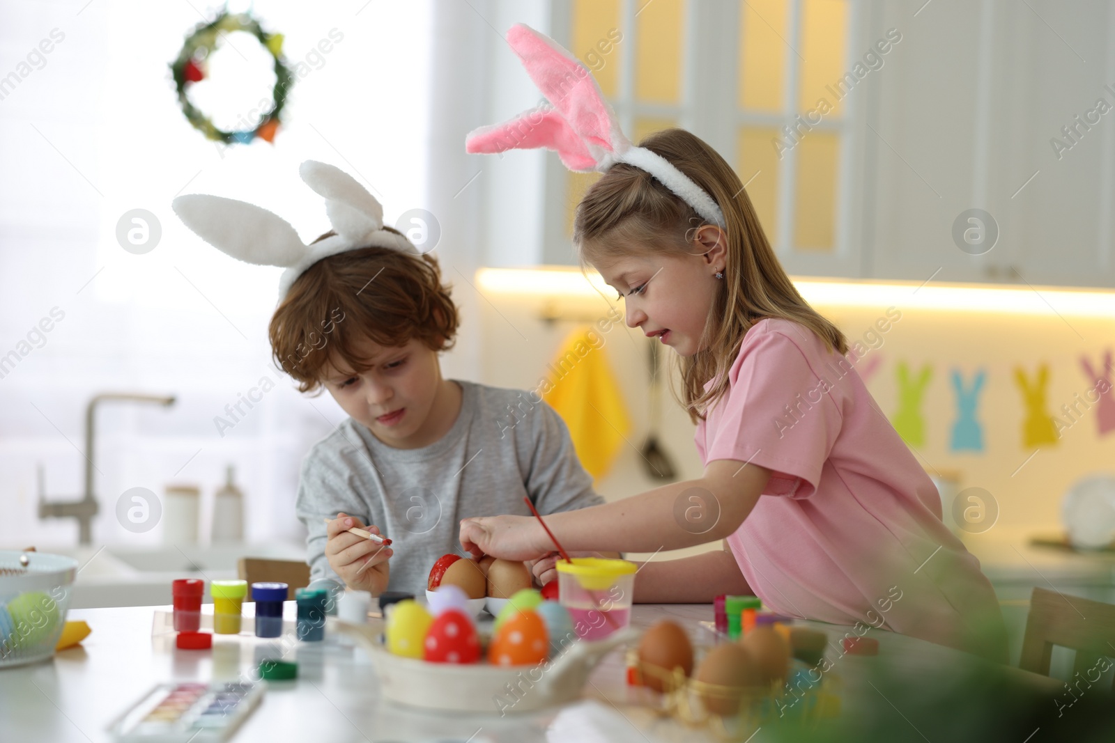 Photo of Easter celebration. Cute children with bunny ears painting eggs at table in kitchen