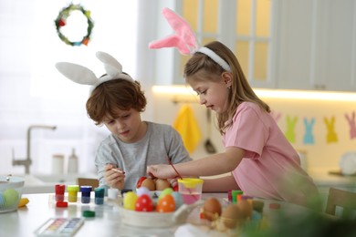 Easter celebration. Cute children with bunny ears painting eggs at table in kitchen