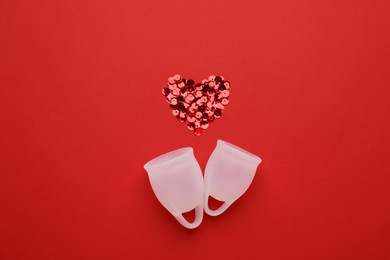Menstrual cups near heart made of sequins on red background, flat lay