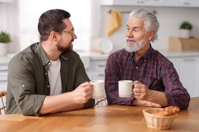 Photo of Happy son and his dad with cups talking in kitchen