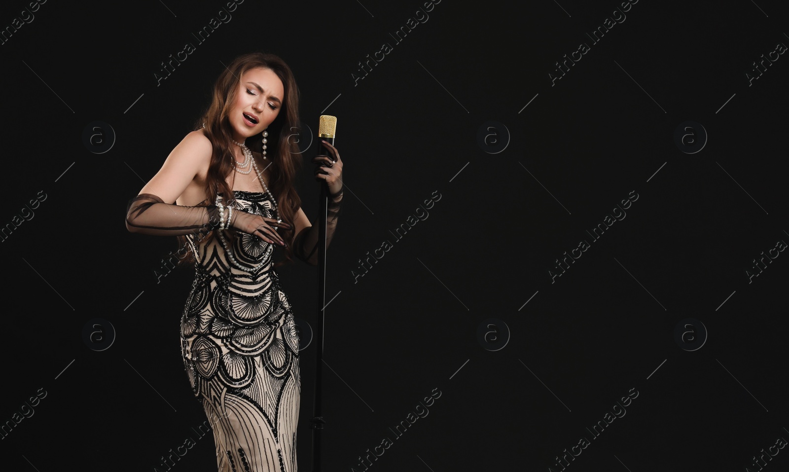 Photo of Beautiful young woman in stylish dress with microphone singing on black background, space for text