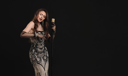 Beautiful young woman in stylish dress with microphone singing on black background, space for text