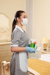Photo of Young chambermaid in mask and gloves holding basket with cleaning products indoors
