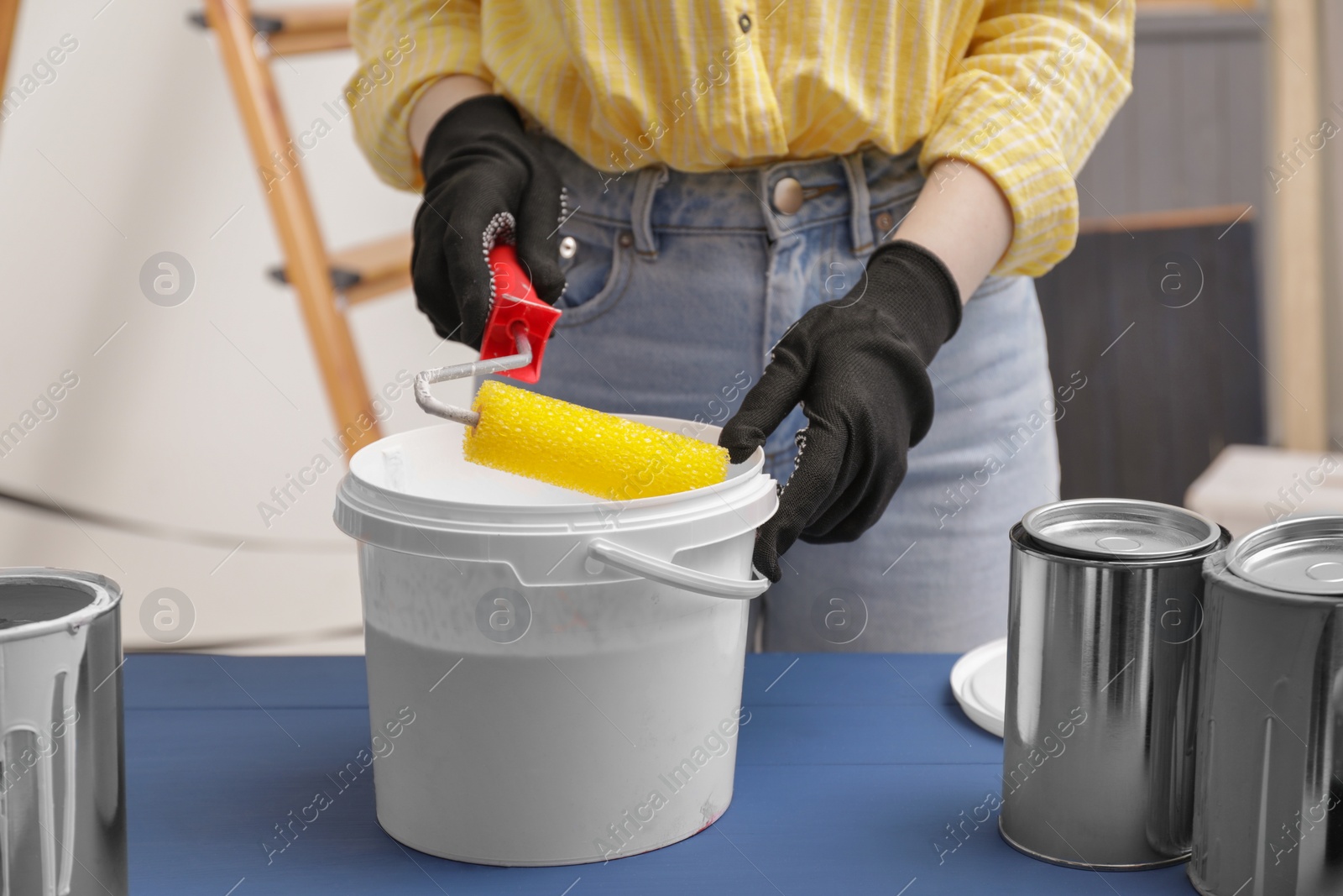 Photo of Woman dipping roller into bucket with paint at blue wooden table indoors, closeup