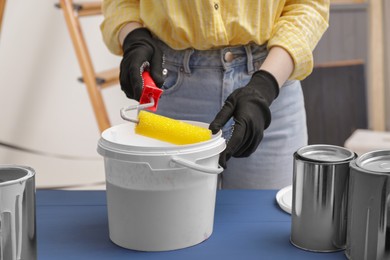 Woman dipping roller into bucket with paint at blue wooden table indoors, closeup