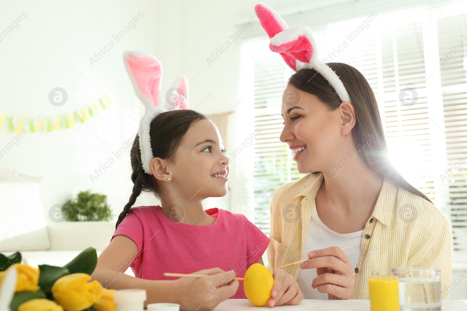 Photo of Happy mother and daughter with bunny ears headbands painting Easter egg at home