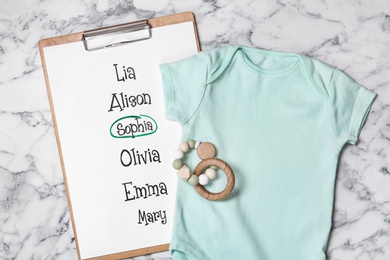 Photo of Bodysuit, list of baby names and toy on white marble background, flat lay