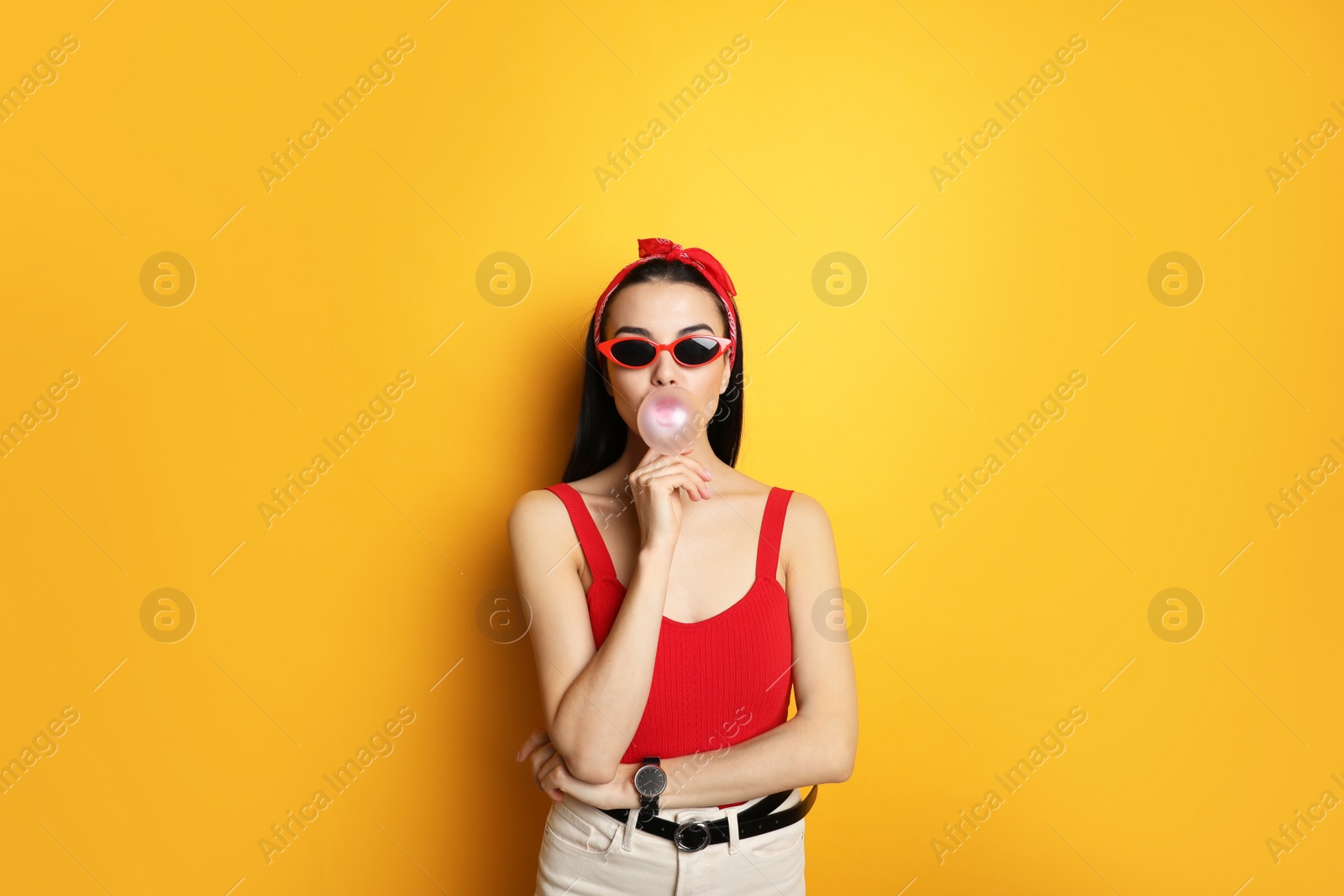 Photo of Fashionable young woman in pin up outfit blowing bubblegum on yellow background