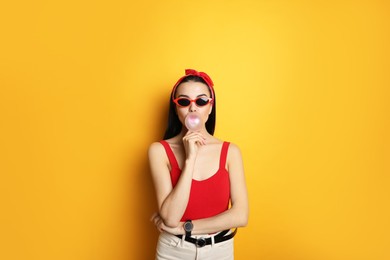Photo of Fashionable young woman in pin up outfit blowing bubblegum on yellow background