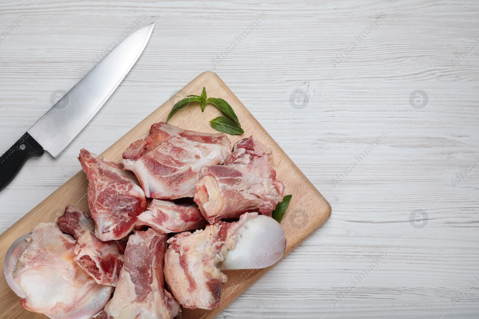 Photo of Cutting board with raw chopped meaty bones, basil and knife on white wooden table, flat lay. Space for text