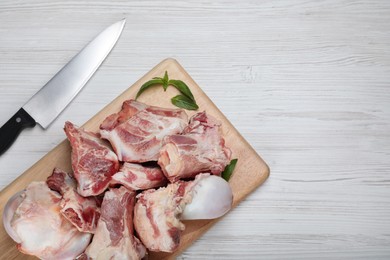 Cutting board with raw chopped meaty bones, basil and knife on white wooden table, flat lay. Space for text