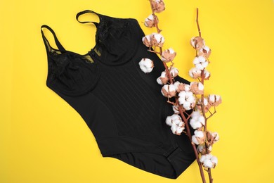 Photo of Elegant black plus size women's underwear and cotton flowers on yellow background, flat lay