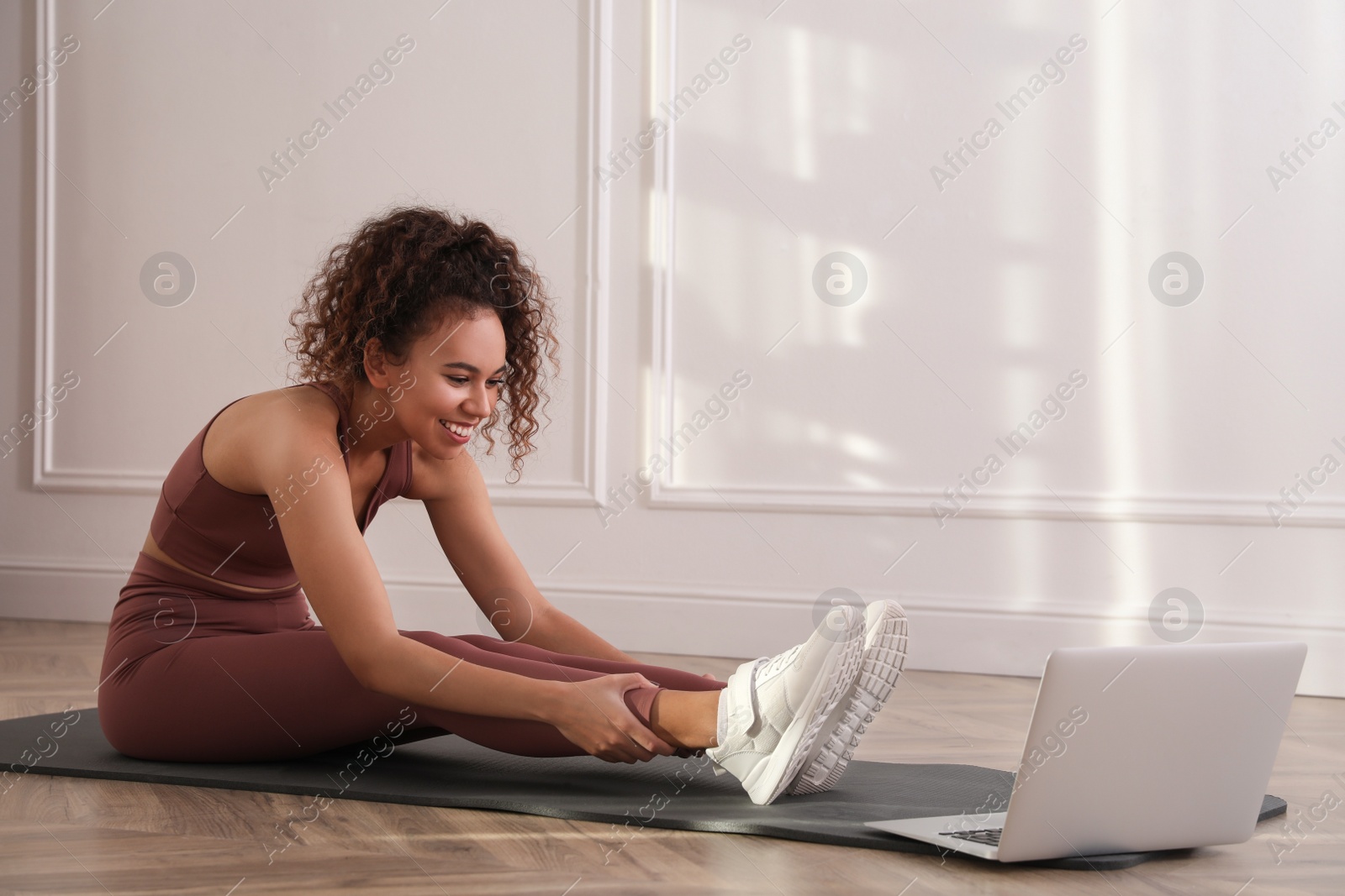 Photo of Beautiful African American woman stretching on yoga mat while watching online class indoors