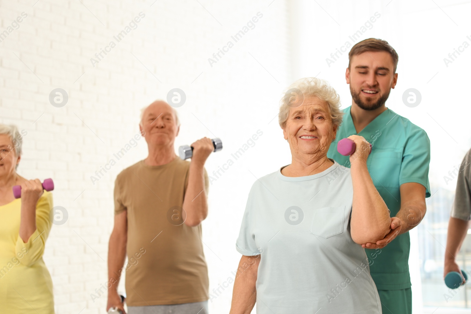 Photo of Care worker helping elderly woman to do exercise with dumbbell in hospital gym