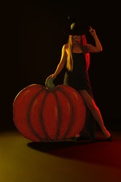 Photo of Young woman wearing witch costume near decorative pumpkin on black background. Halloween party