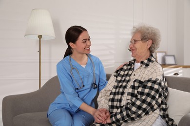 Photo of Young caregiver talking to senior woman on sofa in room. Home health care service