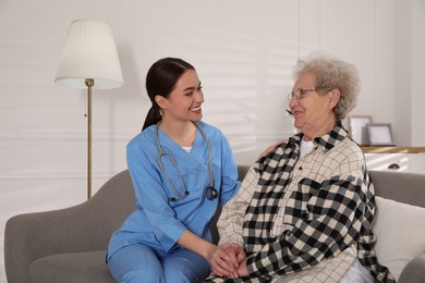 Young caregiver talking to senior woman on sofa in room. Home health care service