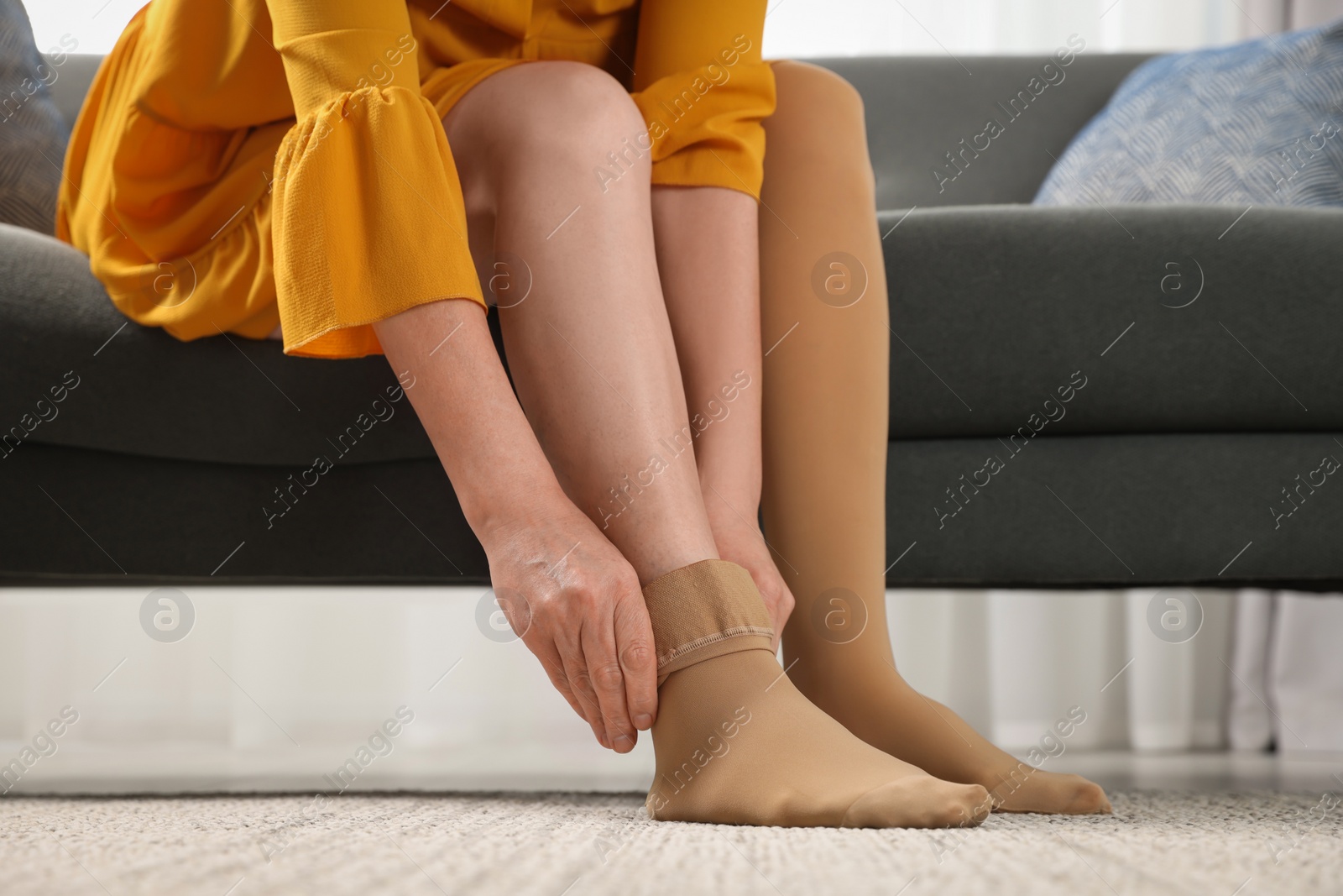 Photo of Woman putting on compression stocking in living room, closeup. Prevention of varicose veins