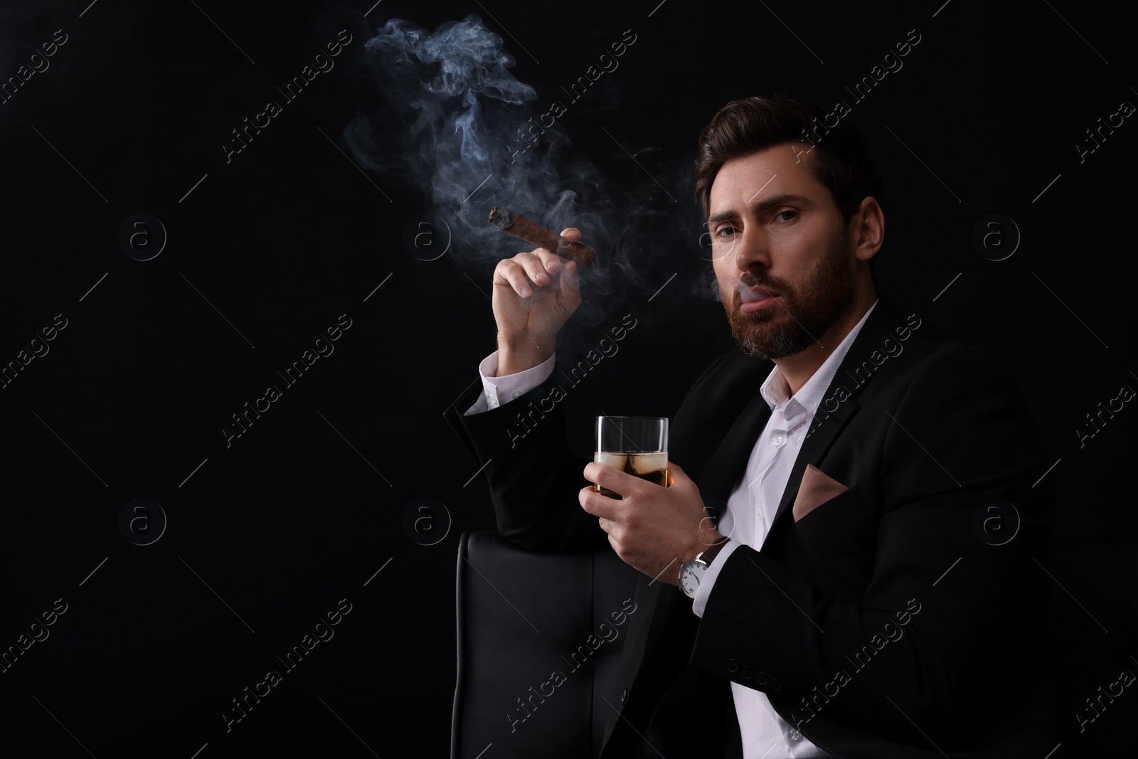 Photo of Handsome man in elegant suit with glass of whiskey smoking cigar on black background. Space for text