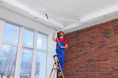 Photo of Handyman painting ceiling on step ladder with roller in room