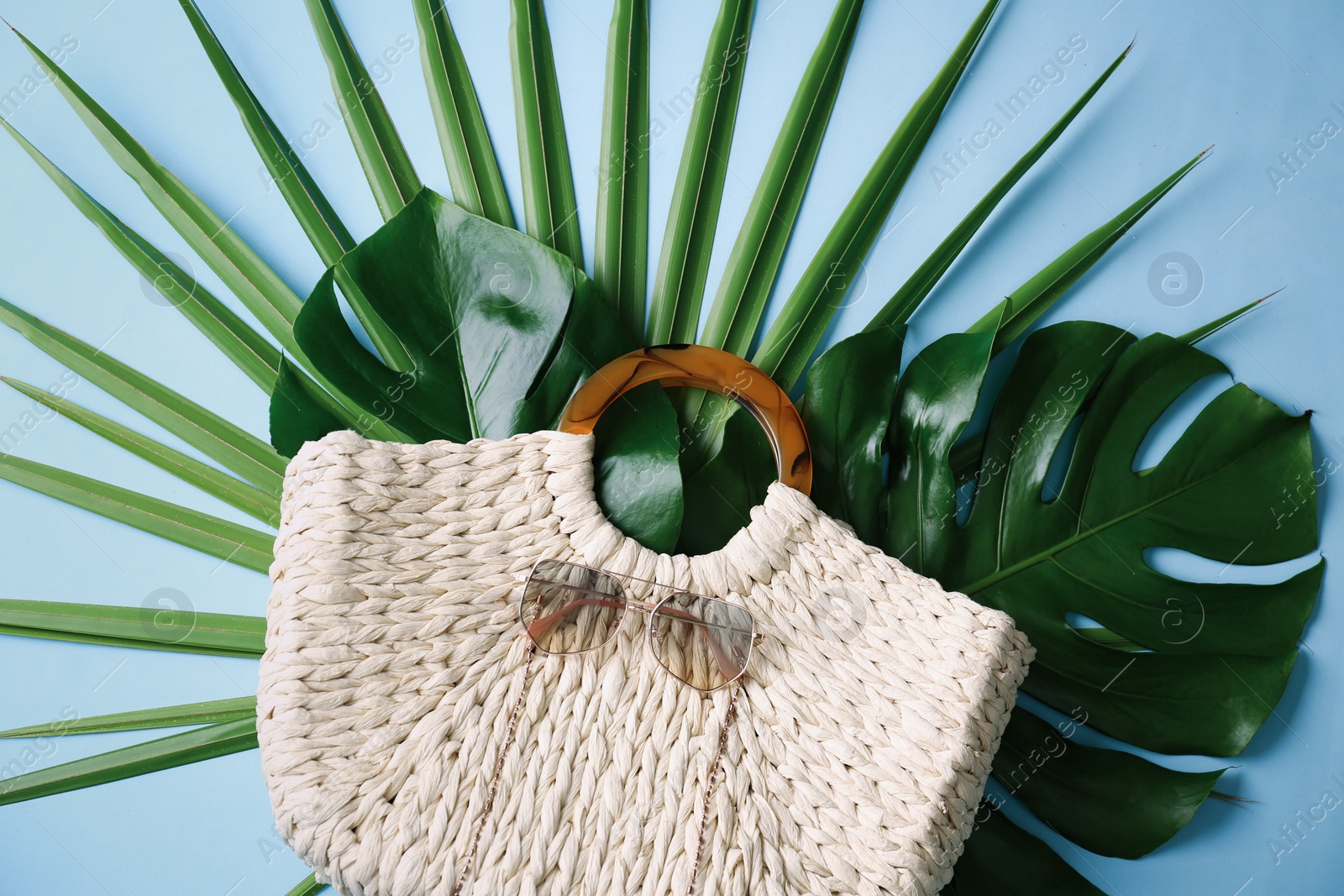 Photo of Elegant woman's straw bag with tropical leaves and sunglasses on light blue background, top view