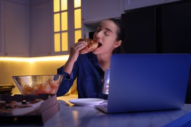 Photo of Young woman eating sandwich while using laptop in kitchen at night. Bad habit