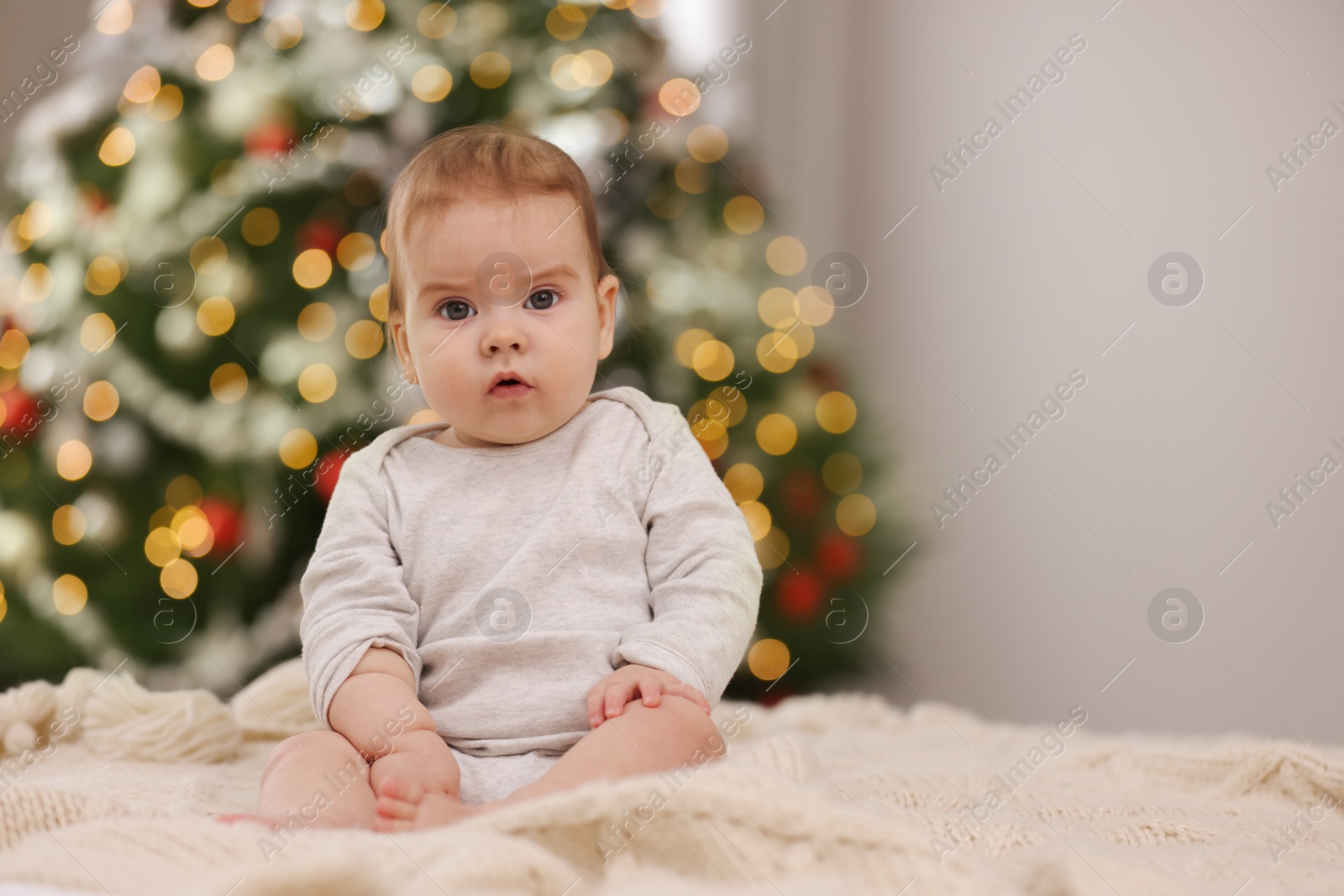 Photo of Cute little baby on knitted blanket against blurred festive lights, space for text. Winter holiday