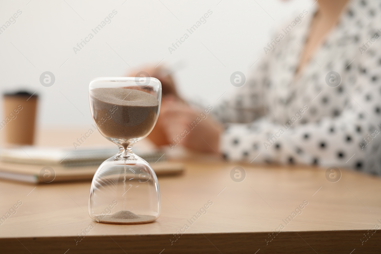 Photo of Hourglass with flowing sand on desk. Woman working indoors, selective focus