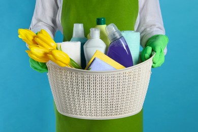 Photo of Spring cleaning. Woman holding basket with detergents, flowers and rags on light blue background, closeup