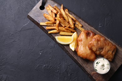 Photo of Tasty fish, chips, lemon and sauce on black table, top view. Space for text