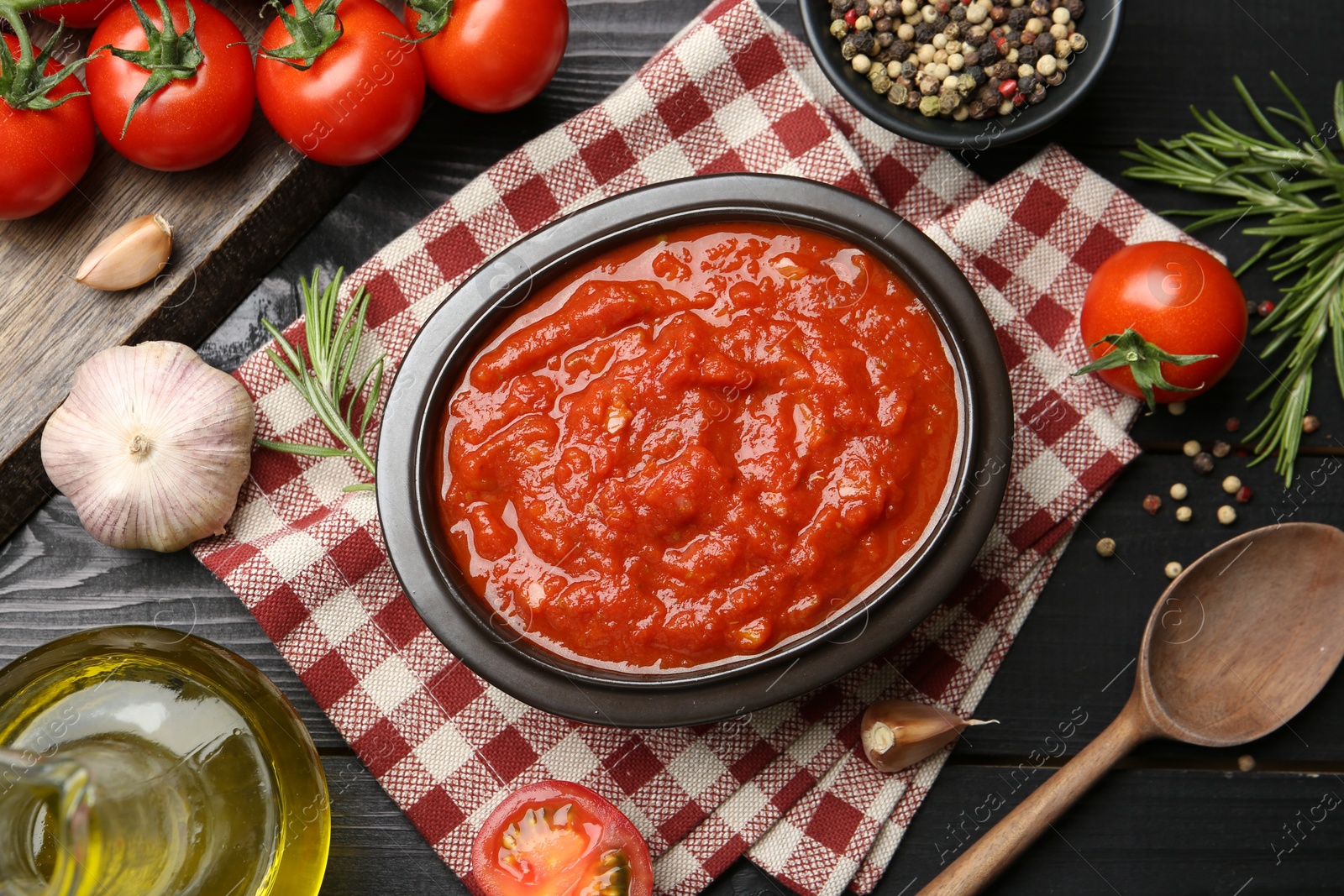 Photo of Homemade tomato sauce in bowl, spoon and fresh ingredients on black wooden table, flat lay