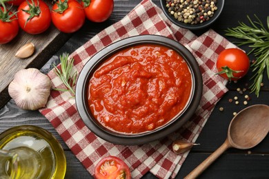 Photo of Homemade tomato sauce in bowl, spoon and fresh ingredients on black wooden table, flat lay