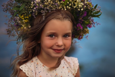 Cute little girl wearing wreath made of beautiful flowers on blurred background, closeup