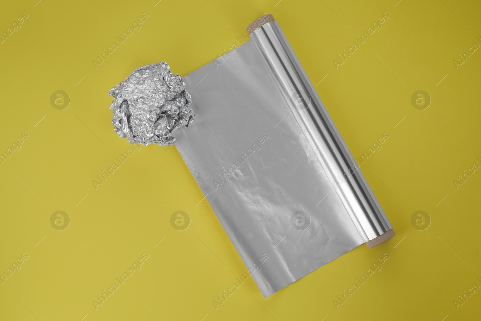Photo of Roll of aluminum foil and crumpled ball on yellow background, top view