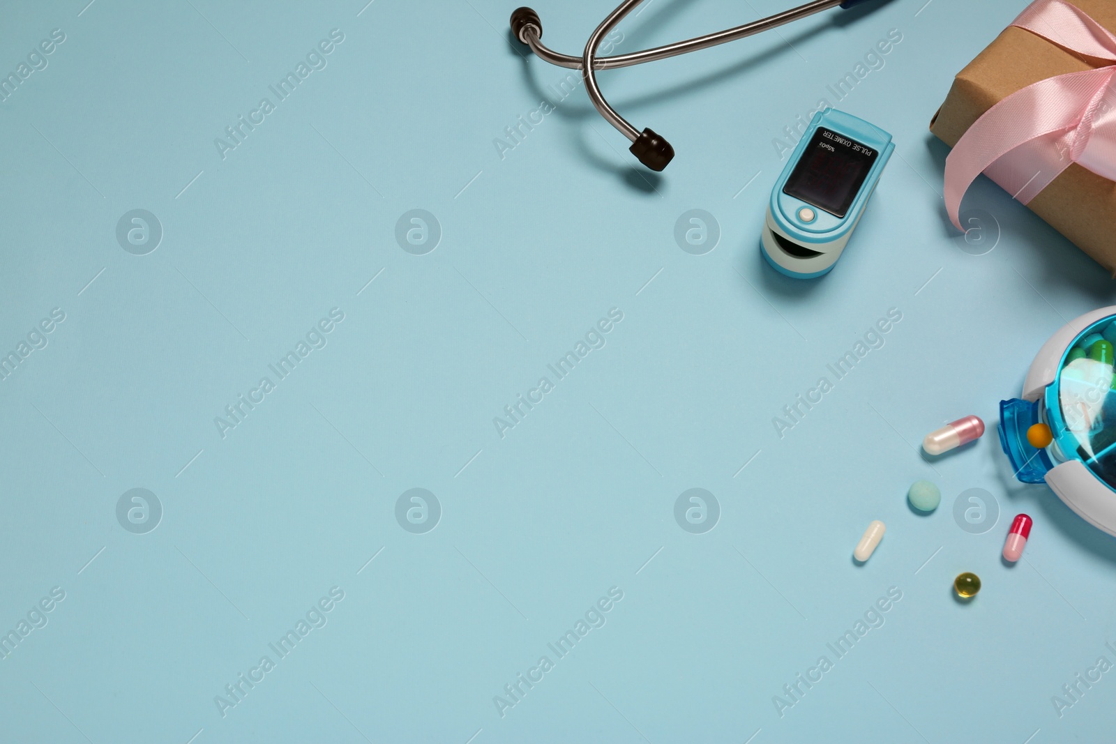 Photo of Stethoscope, pills, box and fingertip pulse oximeter on light blue background, space for text. Medical gift