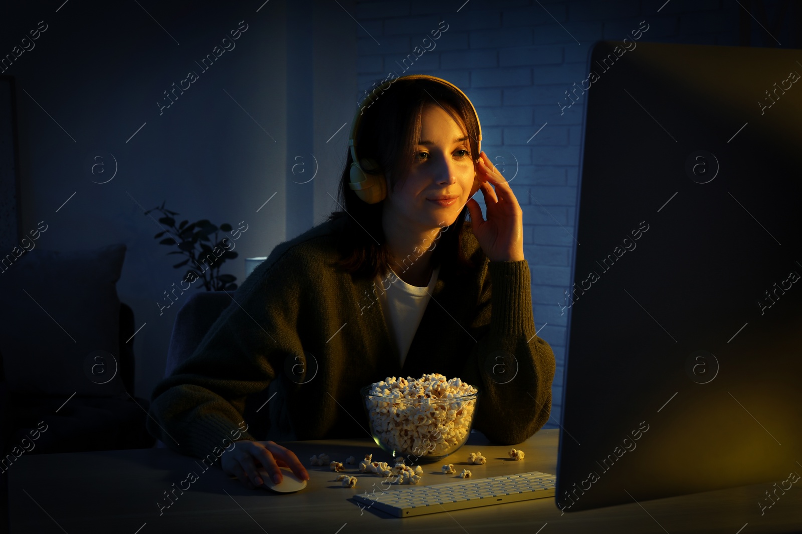 Photo of Beautiful young woman in headphones with popcorn using computer at table indoors at night