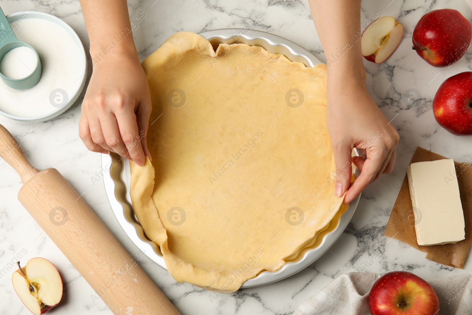 Photo of Woman putting dough for traditional English apple pie into baking dish at white marble table, top view