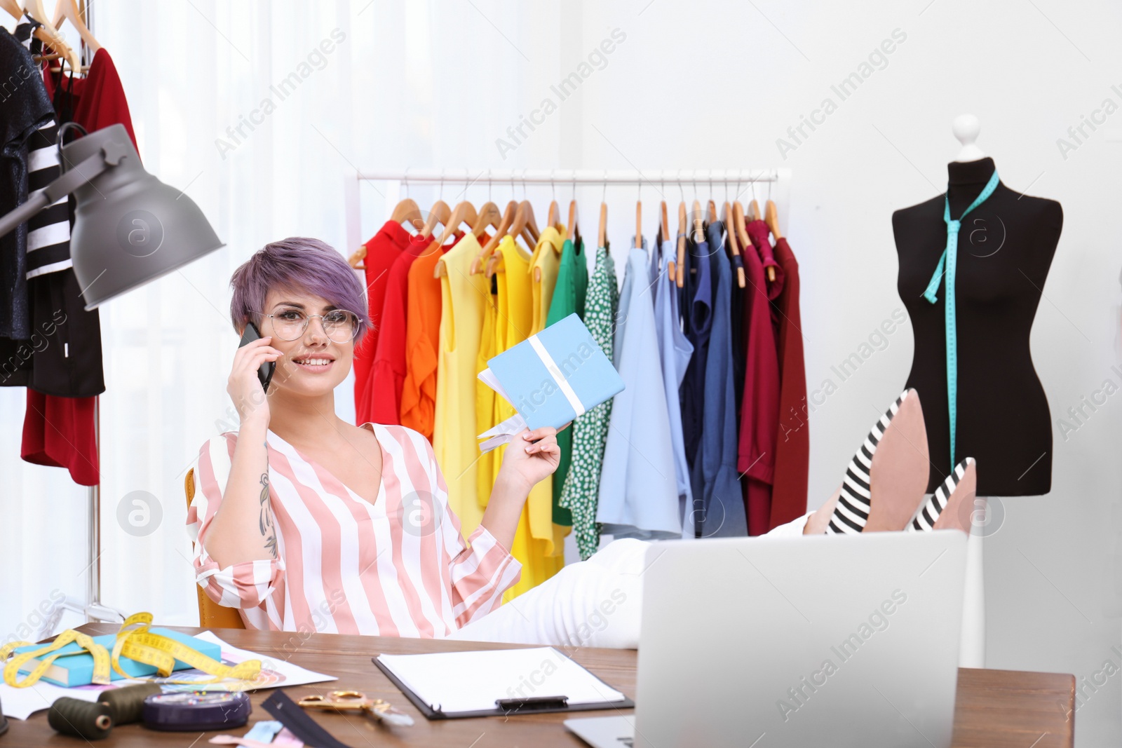 Photo of Beautiful young stylist talking on phone at workplace near rack with clothes