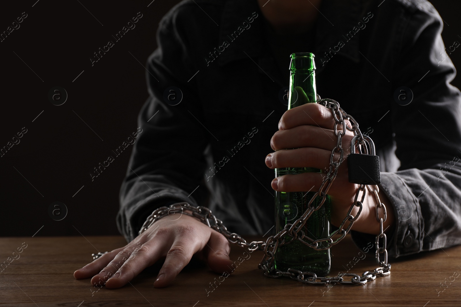 Photo of Alcohol addiction. Man chained with bottle of beer at wooden table, closeup