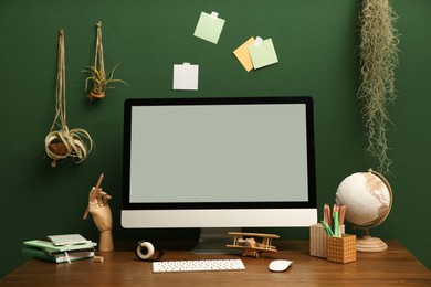 Photo of Stylish workplace with blank computer monitor on wooden desk near green wall. Interior design