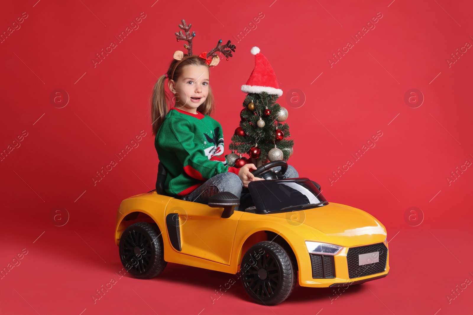 Photo of Cute little girl with Christmas tree driving children's electric toy car on red background