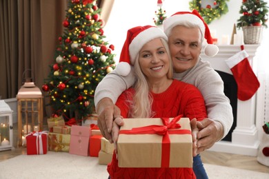 Photo of Happy mature couple in Santa hats with gift box at home, space for text. Christmas celebration
