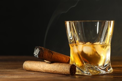 Glass of whiskey with ice cubes and cigars on wooden table, closeup