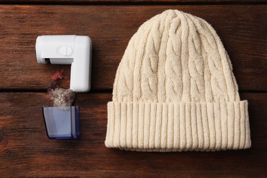 Modern fabric shaver with fuzz and white knitted hat on wooden table, flat lay
