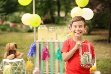Photo of Little boy holding jar with money near lemonade stand in park