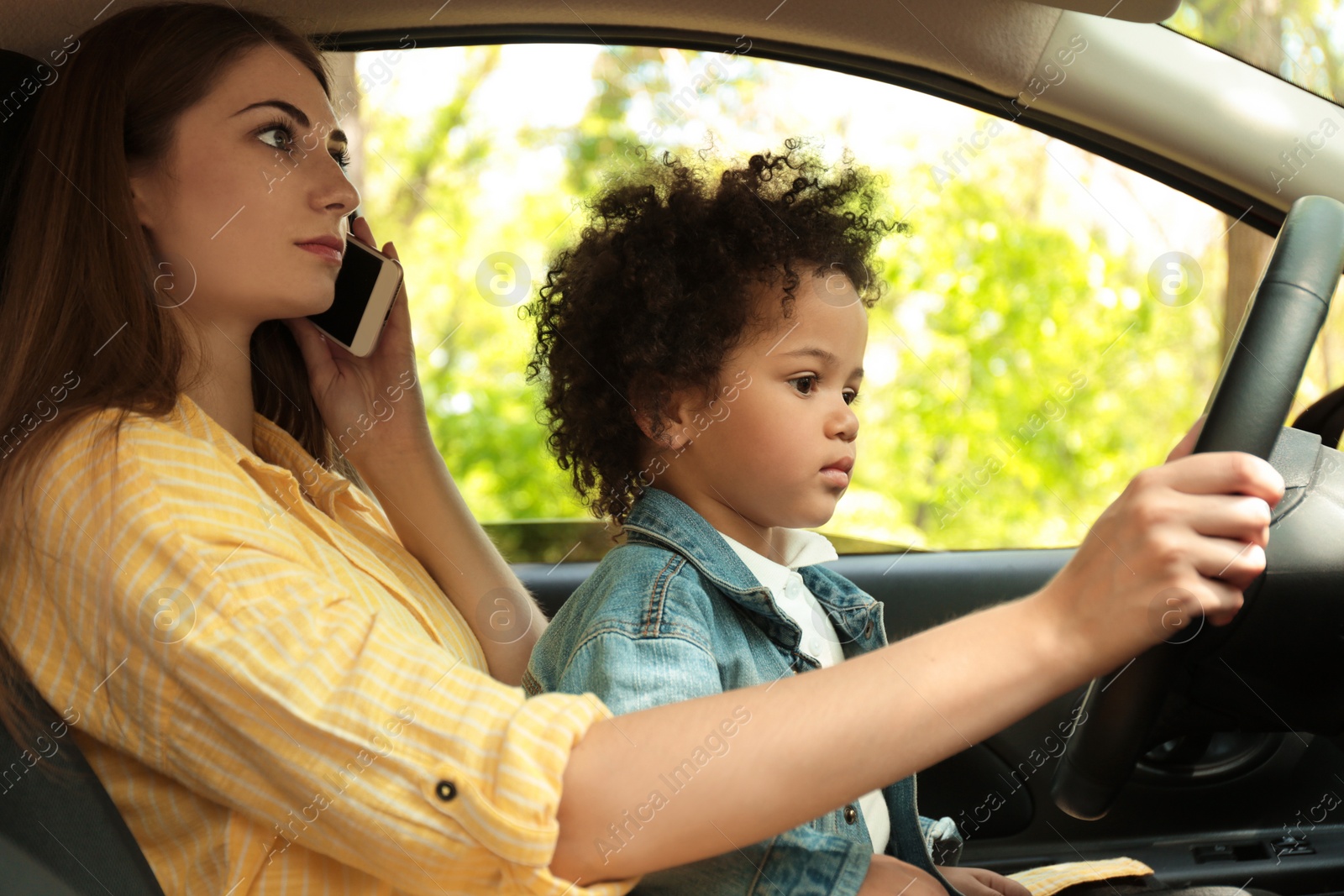 Photo of Mother with little daughter on knees driving car and talking by phone. Child in danger