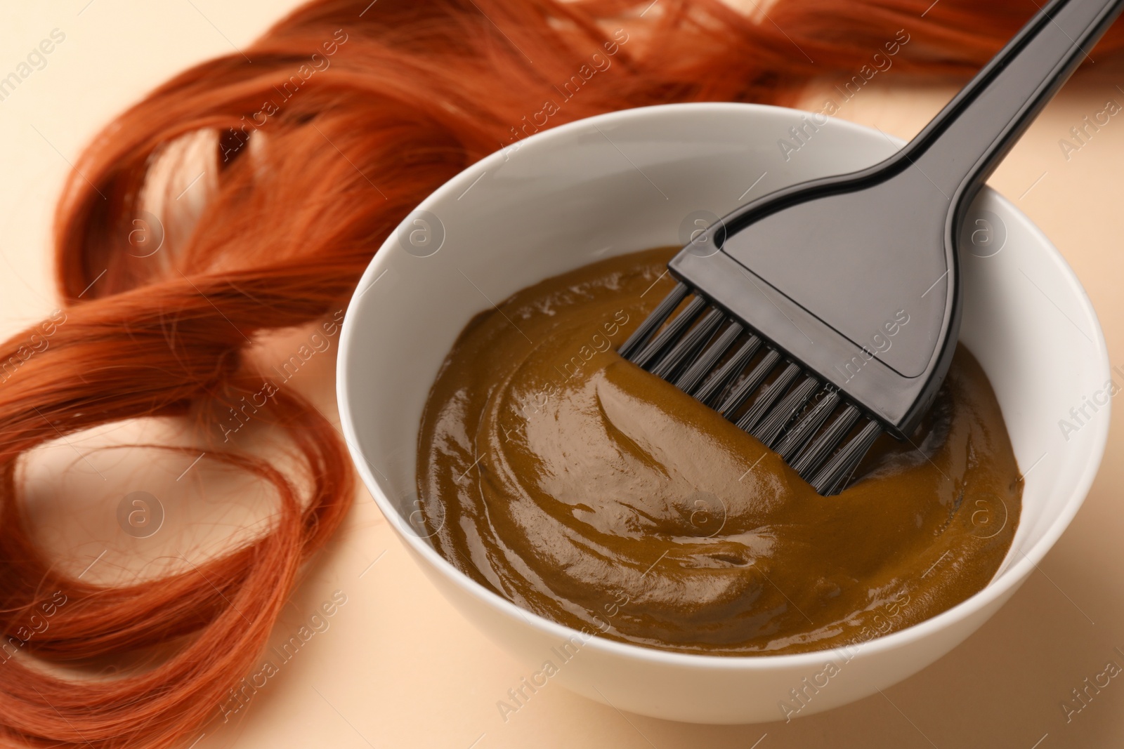 Photo of Bowl of henna cream, brush and red strand on beige background, closeup. Natural hair coloring